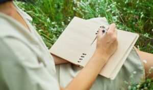 Female woman with pen writing or painting, handwriting on notebook on flower blooming meadow